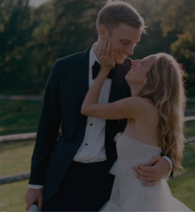 A bride and groom stand outdoors, smiling lovingly at each other. The bride has her hand on the grooms cheek, and they are dressed in wedding attire with greenery and a wooden fence in the background.