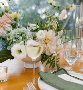 A close-up of an elegant table setting with wine glasses, water glasses, green napkins, a white plate, and a centerpiece of pastel flowers and greenery. A lit candle sits nearby on the wooden table.