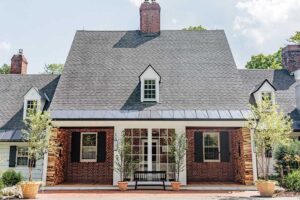 A charming brick house with a steep, dark gray roof, white trim, black shutters, and Vermont luxury hotel accommodations vibes—complete with a central door with glass panes, a black porch bench, and potted plants by the entrance.