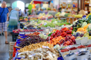 A colorful outdoor market stall displays a variety of fresh fruits and vegetables, including peppers, cherries, apricots, and mushrooms, with shoppers browsing in the background.