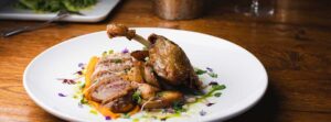 A white plate with roasted duck leg, sliced duck breast, roasted potatoes, carrot strips, microgreens, and edible flowers showcases our Farm to Table Menu Vermont, beautifully served on a wooden table.