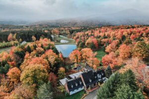 Aerial view of the Edson Hill - Stowe Vermont Inn, a large white house surrounded by vibrant autumn trees, with a pond nearby and rolling hills in the background under a cloudy sky.