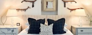 A cozy Edson Hill Guest Room featuring a striped upholstered headboard, black plaid and patterned pillows, two bedside tables with white lamps, plus decorative wooden objects and a mirror on the wall above the bed.