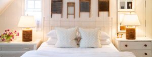A cozy Edson Hill Guest Rooms bedroom features a white bed with patterned pillows, two nightstands with matching lamps, a small clock, and pink flowers in a vase. Vintage washboards hang on the wall above the bed.