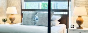 A neatly made bed with multiple gray and white pillows, flanked by two wooden bedside tables. Each table has a lamp with a beige shade and a small decorative item, capturing the inviting charm of Edson Hill Guest Rooms as light streams in through the windows.