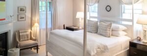 A bright, cozy bedroom at Edson Hill features a white four-poster bed, plaid pillows, two bedside tables with lamps, a soft armchair with a blanket, framed art on white walls, and natural light from two windows with sheer curtains.