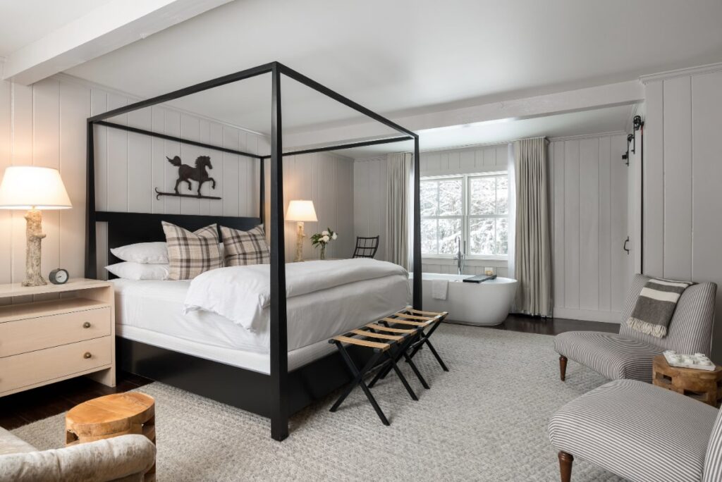 A cozy bedroom with a black four-poster bed, plaid pillows, two striped chairs, and a freestanding bathtub by the window—perfect for relaxing or enjoying special packages. Neutral decor with wood accents and soft lighting complete the inviting space.
