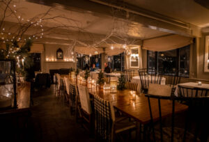A warmly lit restaurant dining room with a long wooden table, surrounded by chairs, candles, string lights, packages, and winter-themed decorations. Three people are seated in conversation at a table in the background.