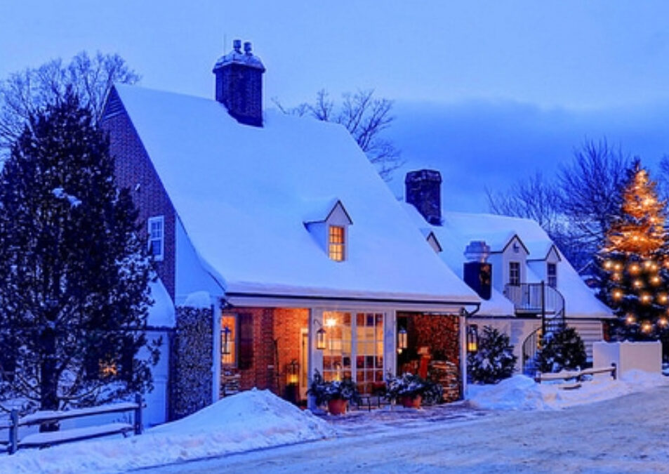 A cozy house covered in snow at dusk, with warm lights glowing from the windows and a decorated Christmas tree outside. Snow blankets the roof and ground, echoing the festive spirit of the 12 Days of Holiday Sales at Edson Hill.