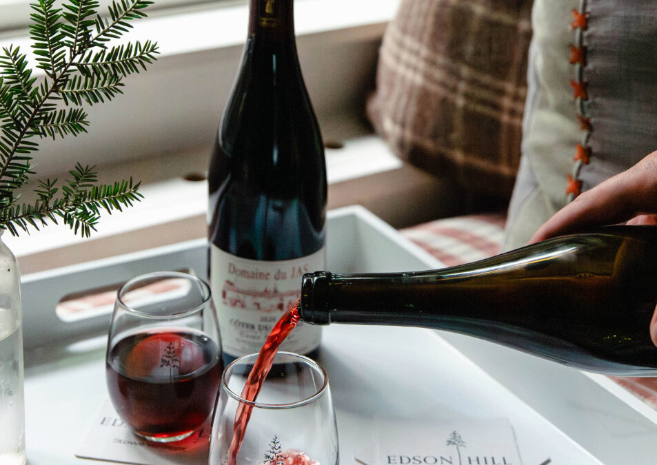 A hand pours red wine into a glass on a white tray with another glass, a wine bottle, and coasters labeled Edson Hill—a cozy scene perfect for savoring the 12 Days of Holiday Sales at Edson Hill. Plaid pillows and a small vase add warmth by the window.