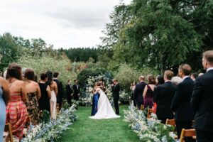 A bride and groom stand together at an outdoor wedding ceremony, surrounded by guests seated on both sides of a flower-lined aisle with lush greenery and trees in the background, making it a perfect setting for customizable wedding packages.