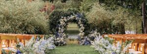 An outdoor weddings setup featuring rows of wooden chairs facing a lush, circular floral arch adorned with white and blue flowers, set against greenery and a serene lakeside backdrop.