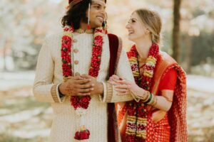 A smiling couple in traditional Indian wedding attire, adorned with red and white flower garlands, stand outdoors at their weddings, holding hands and looking joyfully at each other.