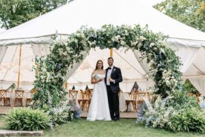 A bride and groom stand smiling under a large floral arch at their weddings celebration in front of a white tent decorated with lights and balloons. The bride wears a white dress, the groom a black tuxedo, surrounded by lush greenery.