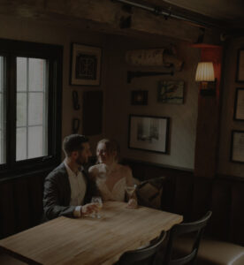 A couple sits closely together at a wooden table in a dimly lit, cozy restaurant, smiling at each other while holding drinks. Framed pictures and soft lighting decorate the wall behind them.
