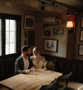 A couple sits closely together at a wooden table in a cozy, dimly lit restaurant, smiling at each other and holding drinks, with framed art and a window in the background.