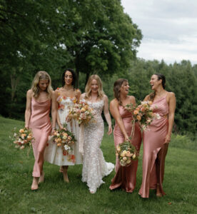 Five women walk outdoors on grass, laughing and smiling. The bride wears a white gown while the four bridesmaids wear matching pink satin dresses and hold bouquets of flowers. Green trees and foliage are in the background.