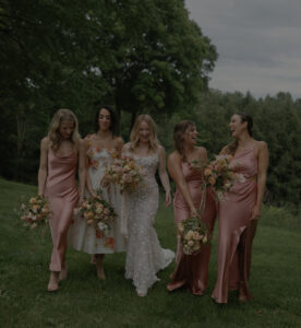 Five women walk together outdoors on grass, holding bouquets. One wears a white wedding dress, while the other four wear matching pink satin dresses. They are smiling and appear joyful, surrounded by lush green trees.