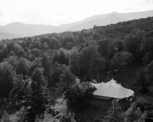A large white event tent is set up in a clearing on a private estate, surrounded by dense forest with distant mountains, perfect for a Private Estate Wedding in Stowe, Vermont. The black and white image captures this serene scene under a partly cloudy sky.