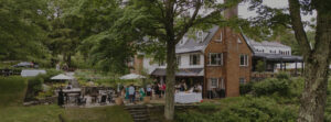 A brick house with a patio is surrounded by trees and greenery. People are gathered outside at tables, some under umbrellas, enjoying a relaxed outdoor event or gathering with special packages available for guests.