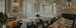 Hanging glass candle holders with lit tealights decorate a cozy, softly lit dining room—perfect for weddings—with elegant table settings and large windows in the background.