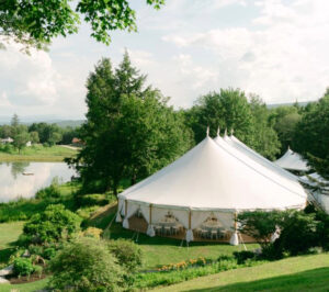 A large white luxury event tent stands on green grass near a pond in Vermont, surrounded by lush trees and gardens under a partly cloudy sky. Tables and chairs are set up inside the tent for an elegant outdoor wedding gathering.