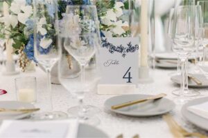 Elegant table setting for weddings with gold cutlery, grey plates, clear wine glasses, flower arrangements, and a “Table 4” card in blue script on a white tablecloth. Candles and white floral decor add a sophisticated touch.