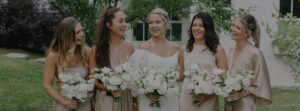 Five women in light-colored dresses, one in a white wedding gown, stand outdoors with white floral bouquets, smiling and laughing together in front of greenery and a white building, celebrating their special day with curated wedding packages.