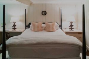 A neatly made bed with white bedding and two pale pink pillows is centered between two modern nightstands, each with a lamp. The room’s white paneled walls and cozy, inviting ambiance evoke the charm of Edson Hill.