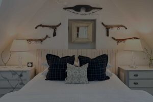 A cozy Edson Hill Guest Rooms bedroom features a bed centered between two nightstands, each with a lamp. Dark plaid pillows and a decorative pillow accent the bed, while vintage items and a framed mirror adorn the sloped wall above.