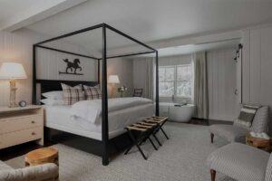 A cozy Edson Hill bedroom with a black four-poster bed, plaid pillows, and a white duvet. A freestanding bathtub sits near the window, accompanied by two striped chairs, a side table with a lamp, and neutral-toned decor.