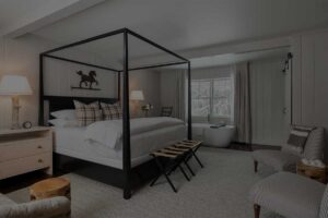 A cozy Edson Hill bedroom with a black four-poster bed, plaid pillows, a beige rug, two cushioned chairs, a small wooden stool, a standing lamp, and a freestanding bathtub near a window with light curtains.