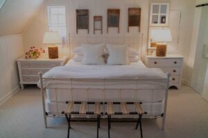 A cozy Edson Hill Guest Rooms bedroom features a white metal bedframe, crisp bedding, two bedside tables with lamps, flowers on the left table, and vintage washboards displayed on the white-paneled wall above the bed.