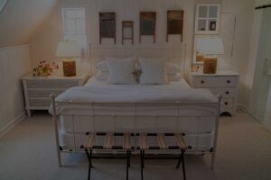 A cozy Edson Hill Guest Room featuring a white metal bed, crisp white bedding, two bedside tables with lamps, flowers on the left table, and vintage washboards hanging on the white paneled wall above the bed.