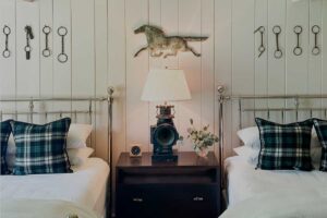 Two twin beds with plaid pillows and white bedding are separated by a dark wooden nightstand holding a lamp, small clock, and greenery. Horse decor and metal horse bits hang on the paneled wall, evoking classic Edson Hill charm.