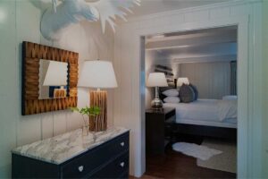 A cozy Edson Hill bedroom features a bed, nightstands, lamps, and a white moose head wall mount. In the foreground, a dresser with a vase of greenery, lamp, and wooden-framed mirror sits against the wall.