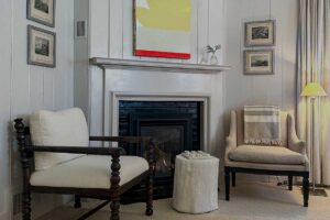 Two armchairs sit by a white fireplace with a modern painting above it, echoing the inviting style of Edson Hill Guest Rooms. A small round table and framed pictures decorate the cozy, softly lit room. A lamp stands near long curtains on the right.