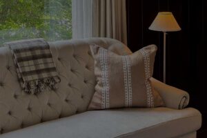 A beige tufted sofa with a plaid blanket and a patterned pillow sits next to a lit floor lamp, evoking Edson Hill charm, with a window revealing green trees in the background.