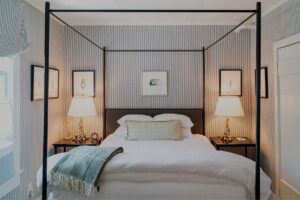 A cozy Edson Hill Guest Rooms bedroom with a four-poster bed, white bedding, a green throw, two bedside tables with lamps, and framed art on striped walls. Light filters in from a window on the left.