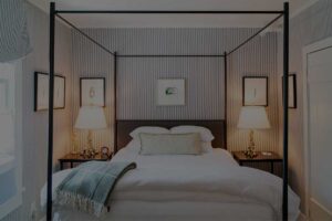 A cozy Edson Hill Guest Rooms bedroom features a four-poster bed with white bedding, a green throw, two bedside tables with lamps, and framed art on grey striped walls. Natural light filters in from the window on the left.