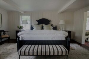 A bright bedroom at Edson Hill features white paneled walls, a large bed with crisp white linens and multiple pillows, two bedside tables with lamps, a striped bench at the foot of the bed, and a cozy rug on the dark floor.