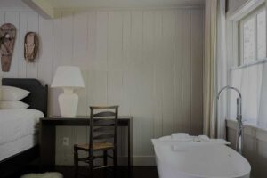 A minimalistic bedroom at Edson Hill features a white freestanding bathtub, a dark wooden desk with a chair, a large white lamp, and a bed with white linens, all set against light-paneled walls and a window with sheer curtains.