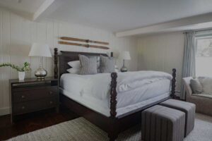 A cozy bedroom at Edson Hill features a dark wooden bed frame, white bedding, patterned pillows, nightstands with lamps, vintage skis mounted on the wall, blue curtains framing the window, and a gray upholstered bench at the foot of the bed.
