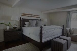 A cozy bedroom at Edson Hill features a dark wooden bed frame, white bedding, and gray accent pillows. Two nightstands with lamps flank the bed, while vintage skis hang above the headboard. A window lets in natural light.