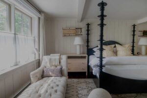 A cozy bedroom at Edson Hill features a black four-poster bed, white bedding, and light wood nightstands. A tufted chaise lounge with a blanket sits by a large window with sheer curtains, letting in soft natural light.