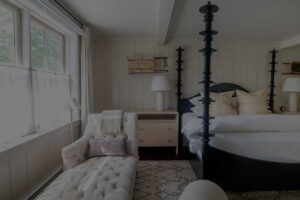 A cozy bedroom reminiscent of Edson Hill with a four-poster bed, a tufted chaise lounge, two bedside tables with lamps, and a window draped in sheer curtains. White walls and soft, neutral decor complete this serene retreat.