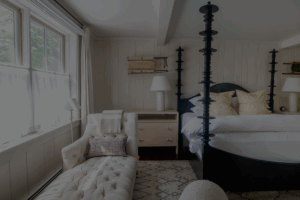 A cozy Edson Hill bedroom with a tall-post bed, white linens, two bedside tables with lamps, a tufted chaise lounge, a patterned rug, and soft natural light from a large window with sheer curtains.