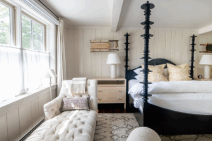 A bright Edson Hill-inspired bedroom with a large window, a white tufted chaise lounge, a wooden nightstand with a lamp, and a bed with tall black posts, white bedding, and patterned pillows. Light décor and neutral tones throughout.