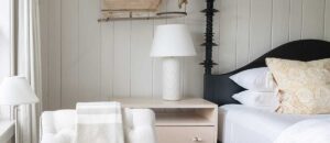 A cozy Edson Hill bedroom with a white chaise lounge, light wood nightstand, white table lamp, and a bed with white linens and patterned yellow pillows against a light paneled wall.