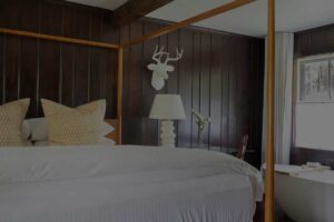 A cozy Edson Hill bedroom with dark wood walls, a four-poster bed topped with white bedding and beige pillows, a lamp on the nightstand, a white deer head wall decoration, and a bathtub near the window.